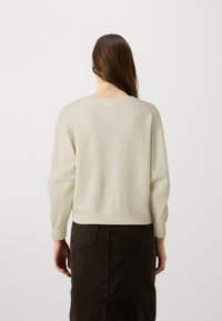 Gai bēšs džemperis ar brīvu piegriezumu, ribinātu apakšmalu un garām piedurknēm, kombinēts ar tumši brūnu svārku ar redzamām šuvju detaļām.