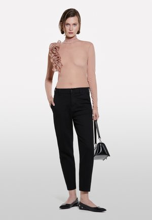 Haut nude à manches longues avec un accent froncé sur une épaule, associé à un pantalon noir ajusté et des chaussures noires plates, plus un petit sac à main noir.