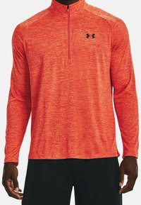 Man wearing a red heathered long-sleeve quarter-zip athletic top with a black logo on the left chest and black pants.