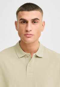 Beige polo skjorte med krage og tre-knappers placket. Stoffet fremstår teksturert og lett, egnet for uformell bruk.