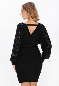 Rochie neagră cu un corp din tricot ajustat și mâneci bufante, cu paiete. Spate în formă de V cu detalii sub formă de curea. Textură netedă în contrast cu mânecile strălucitoare.