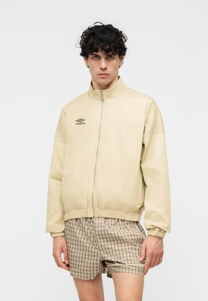 Νέος άνδρας με σγουρά μαύρα μαλλιά, ντυμένος με μπεζ μπουφάν με φερμουάρ και λογότυπο Umbro και μπεζ καρό σορτς, στέκεται μπροστά σε ένα απλό φόντο.