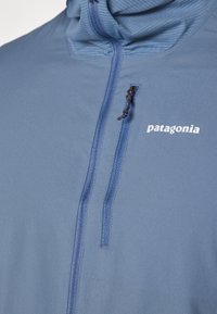 Svetlomodrá mikina Patagonia s polovičným zipsom, textúrovaný materiál, nastaviteľná kapucňa a logo potlačené na hrudi.
