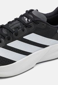 Baskets de sport noires avec une tige en maille, des rayures blanches diagonales, une surface texturée et une semelle en caoutchouc gris clair étiquetée "LIGHTSTRIKE."