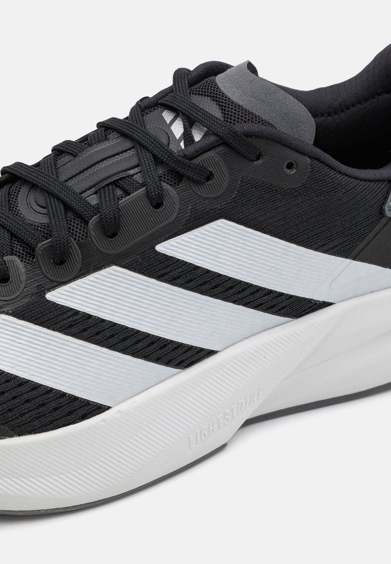 Zapatilla deportiva negra con parte superior de malla, rayas diagonales blancas, superficie texturizada y una suela de goma gris claro etiquetada como "LIGHTSTRIKE".
