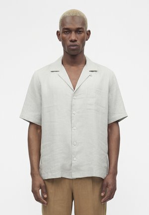 Jeune homme aux cheveux blonds courts portant une chemise grise claire à manches courtes boutonnée et un pantalon marron sur fond blanc uni.