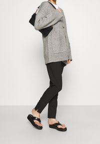 Cardigan en maille grise à coupe oversize, doté de deux poches avant, associé à un pantalon noir slim et des sandales en plastique noires à semelles épaisses.
