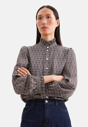 Kvinde med langt mørkt hår bærer en mønstret bluse med høj hals og puffede ærmer samt mørkeblå jeans med høj talje, armene krydset.