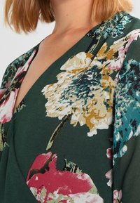 Blusa floral con un fondo verde profundo, que presenta grandes patrones de flores amarillas, rosas y azules. Tela suave, diseño de cuello en V.
