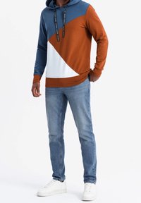 Sudadera con capucha en bloques de color azul, marrón y blanco. Material de mezcla de algodón con cordones y un bolsillo tipo canguro. Combinada con jeans claros.