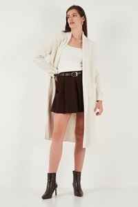 Lichtbeige geribbelde lange cardigan met een sjaalkraag, open voorkant en twee zijzakken, gecombineerd met een witte top en een bruine skort. Bruine enkelhoge laarzen.