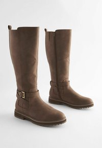 Paire de bottes hautes en cuir marron avec fermeture éclair latérale et bride décorative à la cheville avec boucle, sur fond blanc.