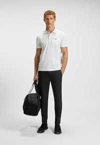Vit piké med subtil textur och svart logotyp, i kombination med svarta slim-fit byxor och svarta sneakers. Modell som håller en svart väska.