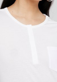 Vit kortärmad topp i mjukt tyg med rund halsringning, en framknäppning med knappar och en bröstficka på vänster sida.