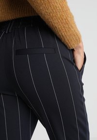 Black pinstriped trousers featuring an elastic waistband and a slit pocket, made from a smooth fabric with a tailored design.
