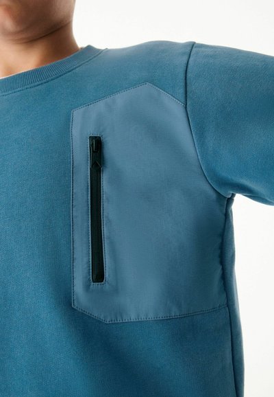 Felpa blu con un tessuto testurizzato, dotata di una tasca opaca a contrasto e chiusura con zip. Il design include una tasca angolata unica.