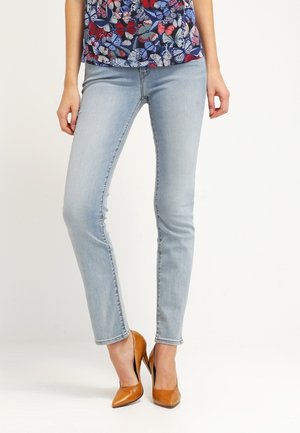 Frau trägt hellblaue Skinny-Jeans, blaues Oberteil mit buntem Schmetterlingsdruck und braune, spitze High Heels, steht vor weißem Hintergrund.