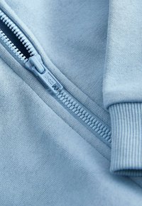 Tejido de sudadera azul claro con una cremallera visible y puños acanalados. Textura suave con detalles de costura sutiles y diseño de cremallera en contraste.