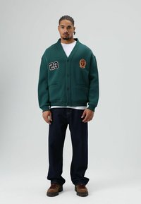 Groen vest met sjaalkraag, knopen en geborduurde patches, gecombineerd met donkere spijkerbroek en bruine veterlaarzen. Gestructureerde stof.