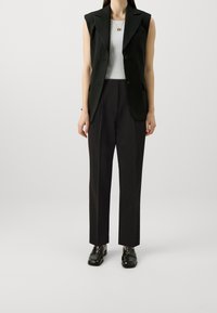 Blazer sans manches noir sur un haut rayé blanc, associé à un pantalon noir et des mocassins noirs brillants. Design simple et ajusté.