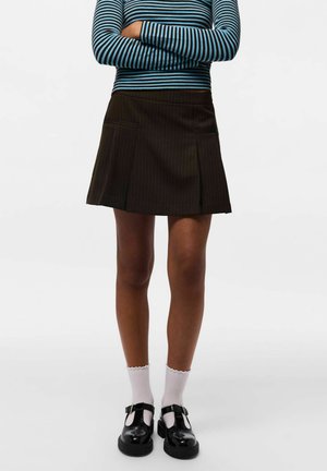 Personne portant une jupe plissée noire, des chaussures Mary Jane noires avec des chaussettes blanches, et un haut à manches longues bleu et noir à rayures, bras croisés.