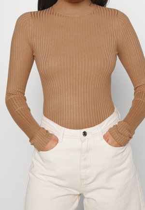 Femme portant un pull à col roulé côtelé couleur camel et un pantalon blanc taille haute, les mains dans les poches.