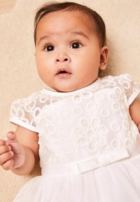 Weißes Kleid mit floralem Stickmuster, kurzen Ärmeln und einer Satin-Schleife. Der Rock besteht aus weichem Tüll und ruht auf einer leichten, strukturierten Oberfläche.