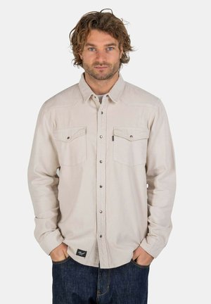 Light beige button-up shirt with two chest pockets, a collar, and long sleeves. Features a subtle texture and dark buttons. Worn with blue jeans.