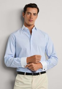 Uomo che indossa una camicia da uomo azzurra chiara con colletto e polsini bianchi, mentre sistema i gemelli, abbinata a pantaloni beige e una cintura nera.