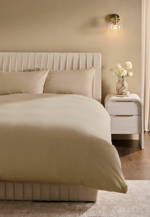 Bett mit beiger Bettwäsche und Kopfteil, Nachttisch mit weißen Rosen in Vase, wandmontierte Lampe, beige Wand und Holzboden.