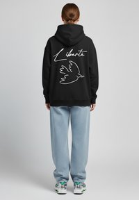 Felpa nera con una silhouette bianca di una colomba e la parola "Liberté" sul retro. Vestibilità ampia abbinata a jeans blu chiaro e sneakers.