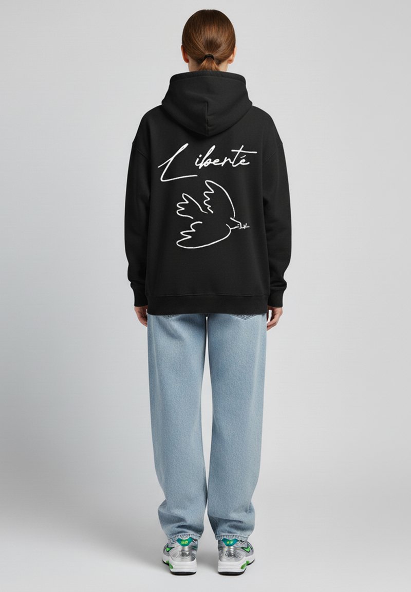 Felpa nera con una silhouette bianca di una colomba e la parola "Liberté" sul retro. Vestibilità ampia abbinata a jeans blu chiaro e sneakers.