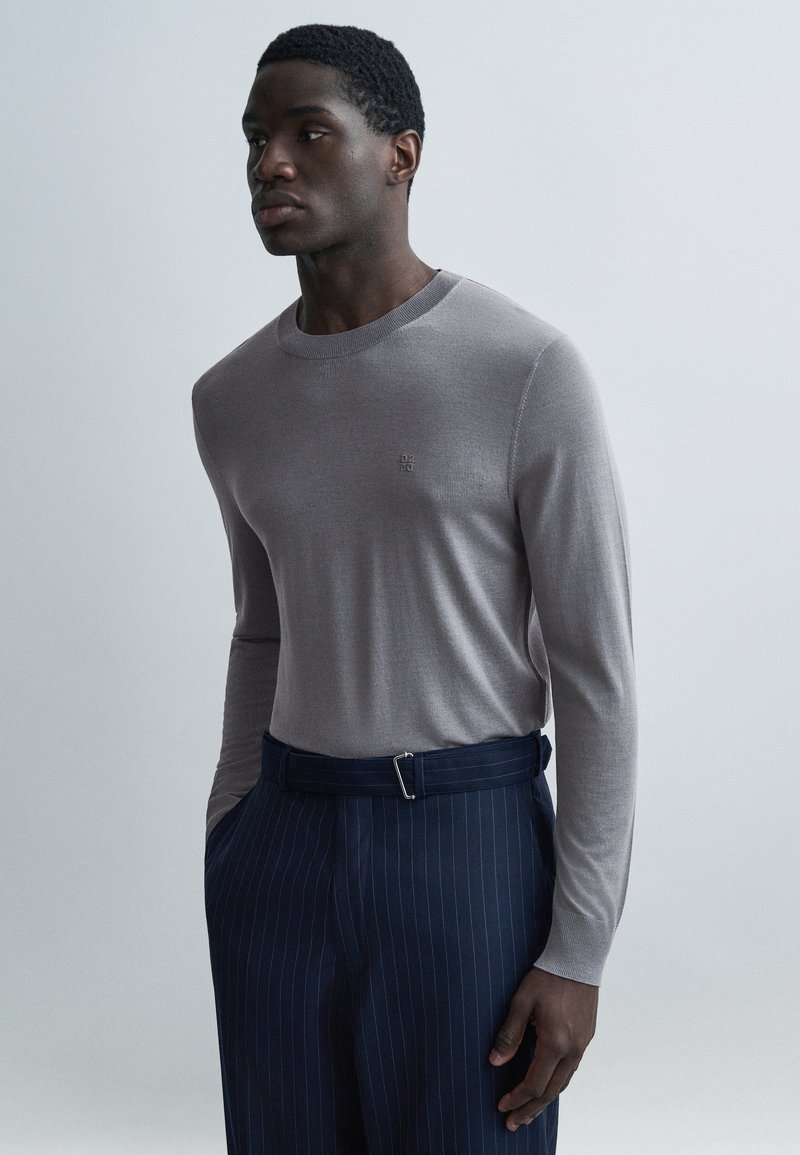 Chemise gris à manches longues en maille avec un col rond, associée à un pantalon rayé bleu marine et une ceinture avec une boucle rectangulaire.