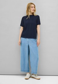 Top de manga corta de textura marina con patrón de olas, combinado con unos vaqueros de pierna ancha en azul claro y zapatillas blancas con acentos en beige.