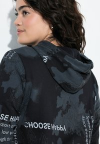 Sudadera negra con un patrón floral texturizado y texto blanco. Tiene una capucha con cordones y un ajuste relajado, mostrando elementos de diseño en capas.