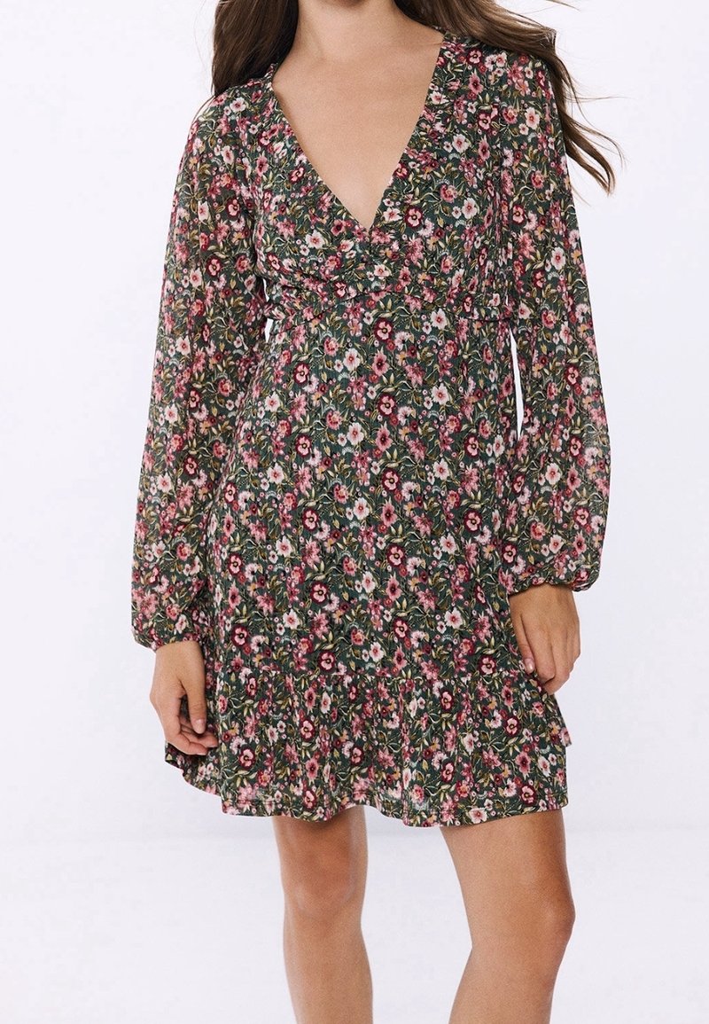 Bloemjurk met een diepe V-hals, lange mouwen en een gelaagd zoom. De stof is lichtgewicht met een groene achtergrond en roze en witte bloemen.