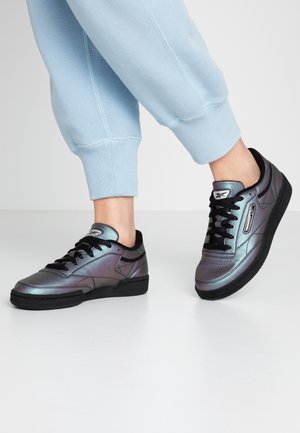 Piernas con pantalones deportivos azul claro que llevan zapatillas metálicas gris-negro brillantes con cordones negros sobre un suelo blanco, un pie ligeramente levantado.