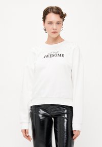 Weißer Sweatshirt mit Perlenakzenten, versehen mit schwarzem Text "wild heart AWESOME", kombiniert mit glänzenden schwarzen Hosen. Insgesamt glatte Textur.