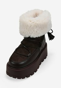 Botín de tobillo de ante sintético marrón con un puño de crema esponjoso, presenta una gruesa suela de plataforma marrón oscuro y cordones decorativos en la parte frontal.