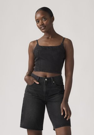 Γυναίκα με σκουρόχρωμο δέρμα που φοράει μαύρο κοντό τοπ χωρίς μανίκια και μαύρα ψηλόμεσα τζιν σορτς, στέκεται με το ένα χέρι στην τσέπη.