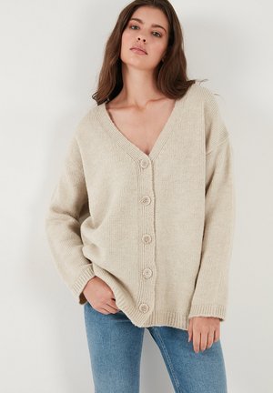 Beige knitted cardigan with a V-neckline, six buttons down the front, and ribbed cuffs. Soft texture, relaxed fit, and oversized design.