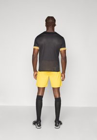 Sort atletisk t-shirt med gule accenter, gule shorts og sorte knæstrømper. Stoffet ser let og åndbart ud.