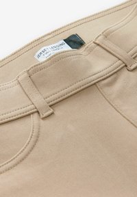 Beige Strumpfhosen aus Jersey mit glatter Textur, ausgestattet mit einem flachen Bund und gesteppten Vorderaschen. Das Etikett trägt die Aufschrift "Jersey Legging Voll Längen".