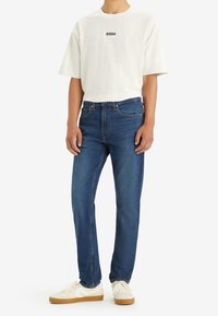 Vit kortärmad skjorta med en svart logotyp, kombinerad med blå denimjeans och vita sneakers med beige sulor. Enkel och avslappnad design.