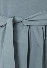 Závojově modré šaty s opaskem vázaným v pase, vyrobené z měkké, hladké látky s plisovanou texturou v dolní části.