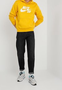 Person som bär en gul Nike SB hoodie, svarta uppviktjeans, vita och blå sneakers, står mot en enkel ljus bakgrund.