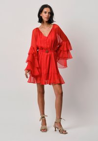 Rotes Chiffonkleid mit Rüschensaum, einem V-Ausschnitt und einer taillierten Taille, die mit einem dekorativen Gürtel verziert ist, kombiniert mit beigen Absatzsandalen.