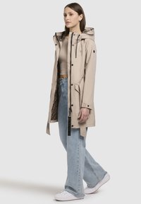 Licht beige waterproof jack met trekkoordcapuchon, binnenvoering met luipaardprint, twee zijzakken en zwarte accenten. Gekozen boven een tan top en blauwe jeans.
