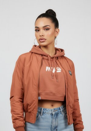 Woman with high bun wearing large hoop earrings, rust-colored cropped hoodie under matching bomber jacket, and light blue jeans against plain background.