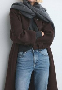 Cappotto di lana marrone indossato sopra un maglione scuro, abbinato a jeans di denim azzurri. Una sciarpa grigia è drappeggiata sciolta intorno al collo.